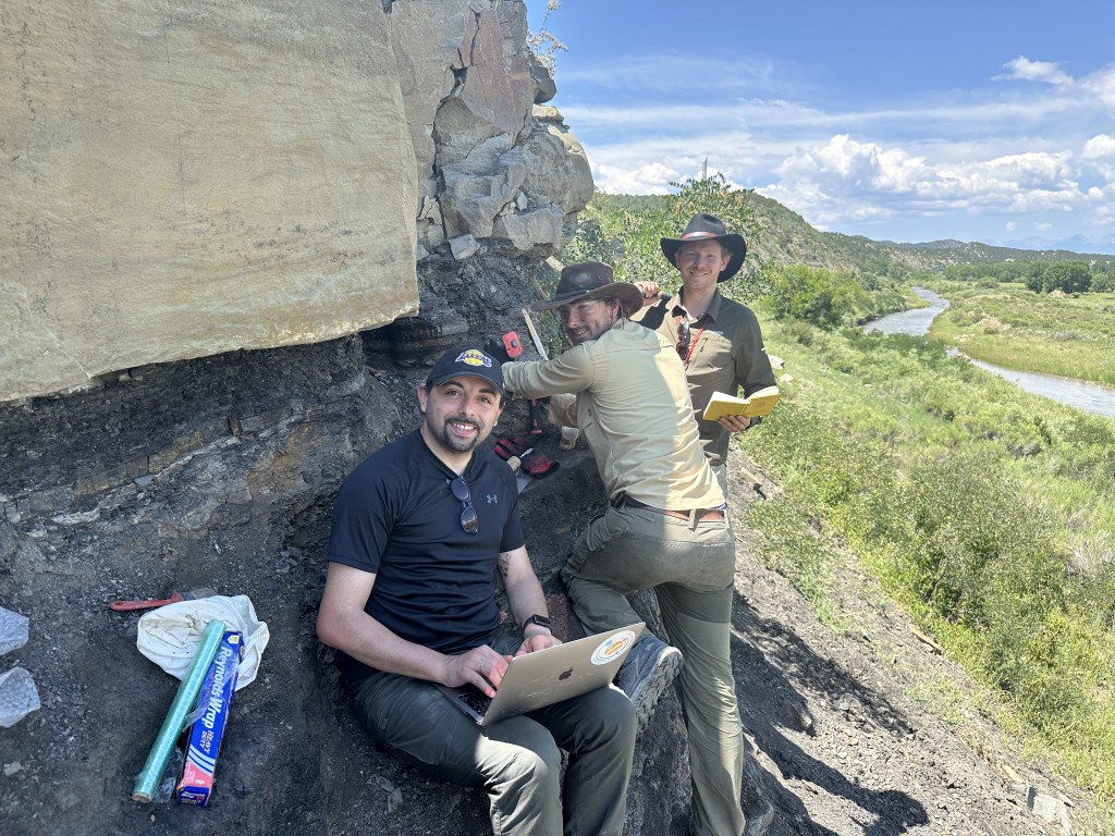 Trois hommes près d'une falaise. L'homme à l'avant est assis avec un ordinateur sur les genoux.
